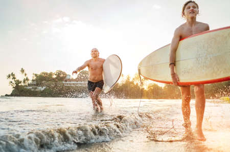 Young teen boy with a surfboard walking with his father by ocean sandy beach after surfing with beautiful sunset background. They are smiling and enjoying their family active vacation.の写真素材