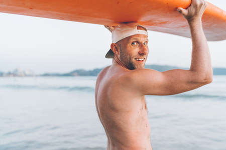 Portrait of shirtless Young Man surfer in baseball cap with a longboard on head smiling at the...