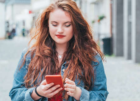 Modern people with technology devices concept image.Vertical portrait of red curled long hair caucasian teen girl walking on the street and browsing the internet using the modern smartphone.の写真素材