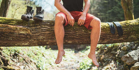 Man sitting on a fallen tree log over the mountain forest river while he waiting for socks nd trekking boots laundry drying. Active people traveling, hiking or trekking concept image.の写真素材