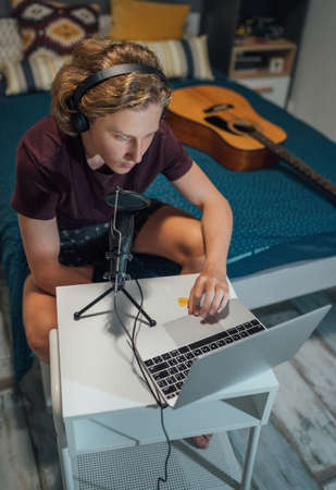 Portrait photo of a young teenage boy in headphones recording voice or chatting with friends using a microphone with pop filter and laptop sitting on a bed in his homeroom.の写真素材