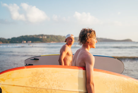 Teen boy with surfboard go to the sea for surfing. He have a winter vacation and enjoying a beautiful sunset light with father on Sri Lanka island. Family active vacation concept.の写真素材