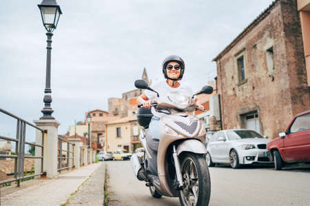 Cheerfully smiling woman in helmet and sunglasses fast riding the moto scooter on the Sicilian old...