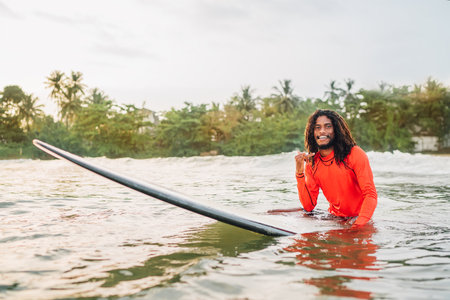 Black long-haired teen man showing shaka sign floating on long surfboard, waiting wave ready surfing...