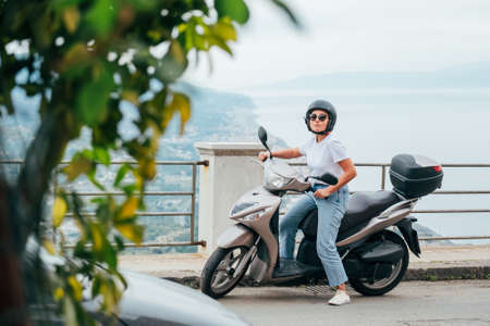 Middle-age woman in helmet and sunglasses on motor scooter on the Sicilian old town streets in the...