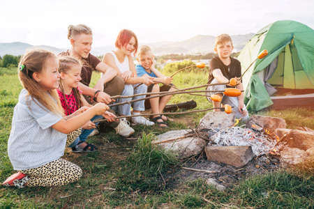 Six kids group Boys and girls cheerfully laughed and roasted sausages on sticks over a campfire flame near the green tent. Outdoor active time spending or camping in Nature concept.の写真素材