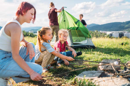 Three sisters smiling and roasting marshmallows and candies on sticks over a campfire flame while two brothers set up the green tent. Happy family outdoor picnic camping activities concept.の写真素材