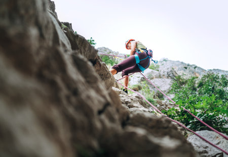 Active climber woman in protective helmet abseiling from cliff rock wall using rope with belay device and climbing harness. Active extreme sports time spending concept.の写真素材