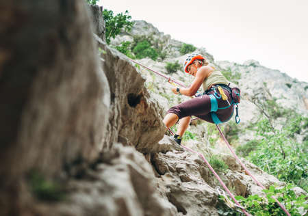 Active climber woman in protective helmet abseiling from cliff rock wall using rope with belay device and climbing harness. Active extreme sports time spending concept.の写真素材