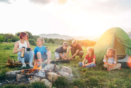Group of smiling kids has a merry conversation near a smoky campfire. They drinking tea , two...