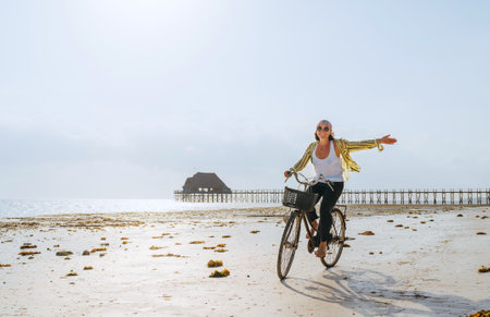 A young female dressed in light summer clothes joyfully threw up her hand while riding old vintage bicycle with a basket on the low tide ocean white sand coast on Kiwengwa beach on Zanzibar, Tansaniaの写真素材