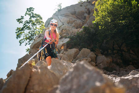 Smiling athletic woman in protective helmet and climbing harness pulling the climbing rope on cliff rock wall in Paklenica National park site in Croatia. Active extreme sports time spending concept.の写真素材