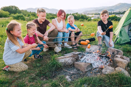 Group of Kids - Boys and girls cheerfully smiling and roasting sausages on long sticks over a campfire flame. Outdoor active time spending or camping in Nature concept.の写真素材