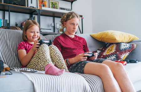 Smiling little Sister with brother sitting on sofa in living room, looking TV and playing console video game using wireless controllers. Careless childhood, modern entertainment technologies concept.の写真素材