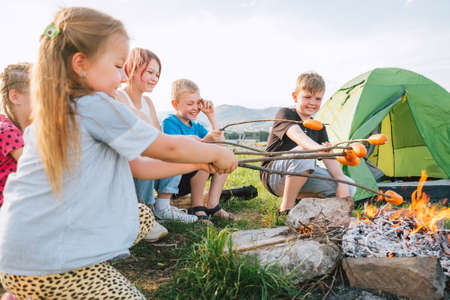Group of Kids - Boys and girls cheerfully smiling and roasting sausages on sticks over a campfire flame near the green tent. Outdoor active time spending or camping in Nature concept.の写真素材