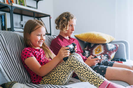 Smiling little Sister with brother sitting on sofa in living room, looking TV and playing console video game using wireless controllers. Careless childhood, modern entertainment technologies concept.の写真素材
