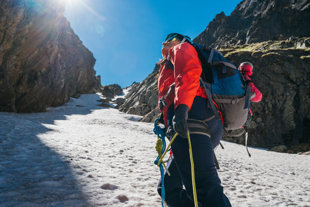 Team roping up woman dressed high altitude mountaineering clothes and harness climbing with backpack by snowy slope in the couloir with backlight sun on blue sky. Vysoke Tatry,Slovの写真素材