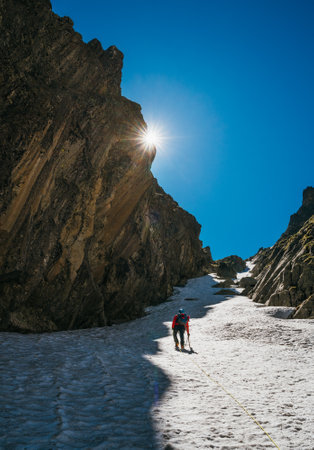 Team roping up woman with climbing axe dressed high altitude mountaineering clothes with backpack walking by snowy slope in the couloir with backlight sun on blue sky.の写真素材