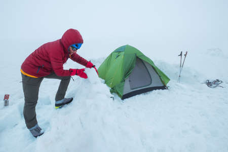 Smiling man dressed red warm mountaineering down jacket making the protective wall from snow bricks...
