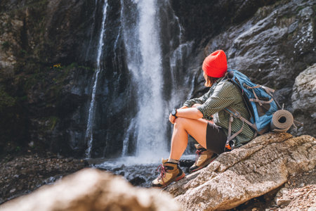 Woman with a backpack in red hat dressed in active trekking clothes sitting near the mountain river waterfall and enjoying the splashing Nature power. Traveling, trekking, and a nature concept image.の写真素材