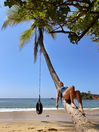 Female dressed blue swim suite lying and relaxing on the coconut palm tree trunk with hanging swing wheel and suntanning on the sandy Indian ocean beach, Mirissa district^ Sri Lanka.の写真素材