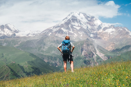 Backpacker woman with a backpack standing and enjoying snowy slopes of Kazbek 5054m mountain while she walking by green grass hill. East Caucasus mountains, Georgia.の写真素材