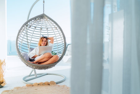 Beautiful middle-aged woman sitting in a comfortable hanging chair on the open house terrace with a modern laptop and browsing in internet doing shopping online. Modern technology and home lifestyle.の写真素材