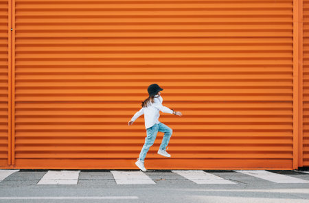Little fashion-dressed girl funny jumping crossing a pedestrian zebra crosswalk on the orange wall...