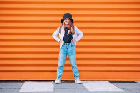 Little fashion-dressed girl portrait sincerely smiling while looking at camera on a pedestrian zebra crosswalk on orange wall background. Urban people living and a street everyday life concept imageの写真素材