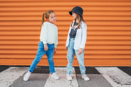Two little girls sisters dressed kids fashion vogue style clothes posing at camera on the city pedestrian asphalt road near the orange wall background. Happy modern childhood concept image.の写真素材