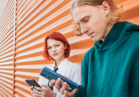 Young caucasian teen girl portrait gazing at camera. She browsing smartphone devices with a friend boy standing near orange wall and. Careless young teenhood time and a modern technology concept imageの写真素材