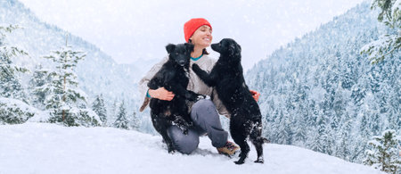 A young woman in warm clothes walking her 2 dogs in a picturesque snowy mountain outdoor. Female laughing and playing with pets and one dog licking an owner's cheek.Human and pets winter concept imageの写真素材