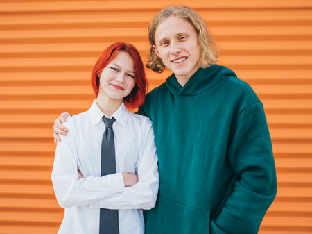 Portraits of two smiling caucasian teen friends long haired boy and dyed hair girl posing for photo while they standing near the orange wall background. Careless young teenhood . time concept image.の写真素材