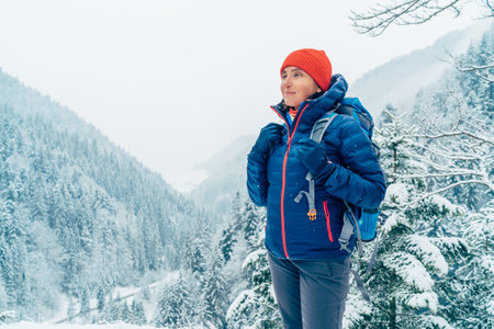 Female backpacker with backpack dressed warm down jacket enjoying snowy mountains landscape while she trekking winter mountain forest route. Active people in the nature concept image.の写真素材