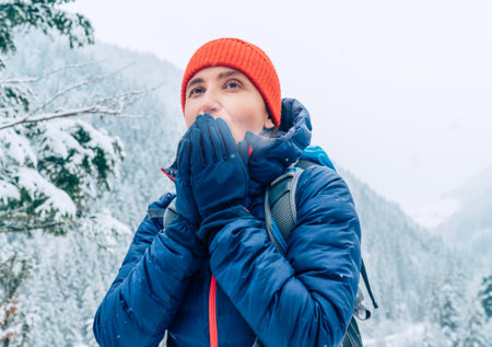 Female backpacker with backpack dressed warm down jacket warming palm hands and enjoying snowy mountains landscape while she trekking winter mountain forest route. Active people in nature concept.の写真素材