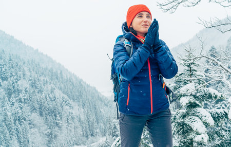 Female backpacker with backpack dressed warm down jacket warming palm hands and enjoying snowy mountains landscape while she trekking winter mountain forest route. Active people in nature concept.の写真素材