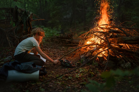 Young caucasian teenage boy adding dry wood to big campfire in the evening time. Camping, traveling...