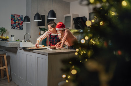 Cute little girl in red Santa hat with mother making homemade Christmas gingerbread cookies using cookie cutters together in home kitchen. Happy family holidays preparation and childhood concept.の写真素材