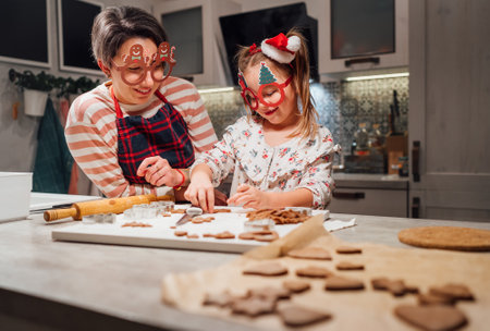 Cute little girl with mother in funny glasses making homemade dough Christmas gingerbread cookies using cookie cutters together in home kitchen. Happy family holidays preparation, childhood concept.の写真素材