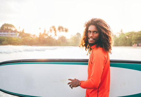 Portrait of black long-haired teen boy with surfboard ready for surfing with sunset backlight. He walking in Indian ocean waves. Extreme water sports and exotic countries concept, Udawalawe, Sri Lankaの写真素材