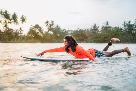 Black long-haired man paddling on long surfboard to the surfing spot in Indian ocean. Palm grove litted sunset rays in the background. Extreme water sports and traveling to exotic countries concept.の写真素材
