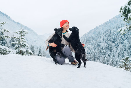 A young woman in warm clothes walking her 2 dogs in a picturesque snowy mountain outdoor. Female laughing and playing with pets and one dog licking an owner's cheek.Human and pets winter concept imageの写真素材