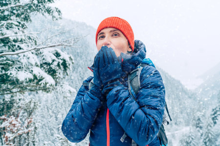 Female backpacker with backpack dressed warm down jacket warming palm hands and enjoying snowy...