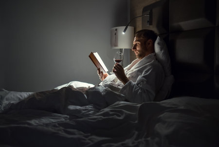 Midle-aged man relaxing rin bed reading book holding a glass of red wine with bedside lamp turned on. Evening relaxation, hobbies, free time concept. Adulthood concept.の写真素材