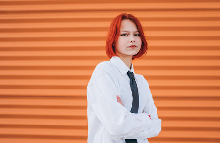 Bright portrait of teenage girl with painted red dyed hair in white school shirt and necktie standing near orange wall background with hands crossed body posture.の写真素材
