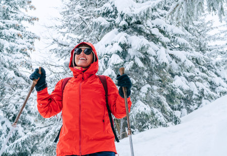 Smiling female trekker dressed red jacket in sunglases with trekking poles walking by snowy slope with fir-trees covered snow, Low Tatra mountains, Slovakia. Beauty in Nature and active people conceptの写真素材