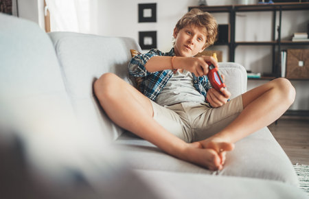 Teenager boy sitting on living room sofa and enthusiastically playing game console using gamepad. Carefree childhood time concept image.の写真素材
