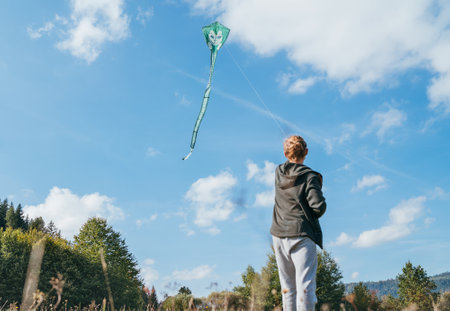 A young teenage boy with a flying green color kite toy on the high grass meadow in the mountain fields. Happy teenhood moments or outdoor time spending concept image.の写真素材