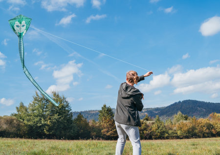 A young teenage boy with a flying green color kite toy on the high grass meadow in the mountain fields. Happy teenhood moments or outdoor time spending concept image.の写真素材