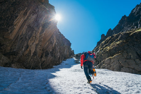 Team roping up woman dressed high altitude mountaineering clothes and harness climbing with backpack by snowy slope in the couloir with backlight sun on blue sky. Vysoke Tatry,Slovakia.の写真素材